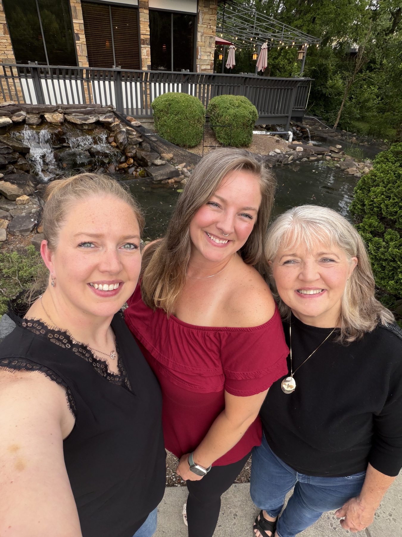 Aleasha in a red top standing between two other women beside a garden pond and waterfall.